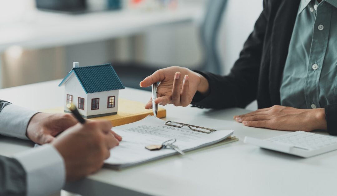 Two people in blazers sit at a desk with paperwork, keys, and a small model house between them. One signs a document.