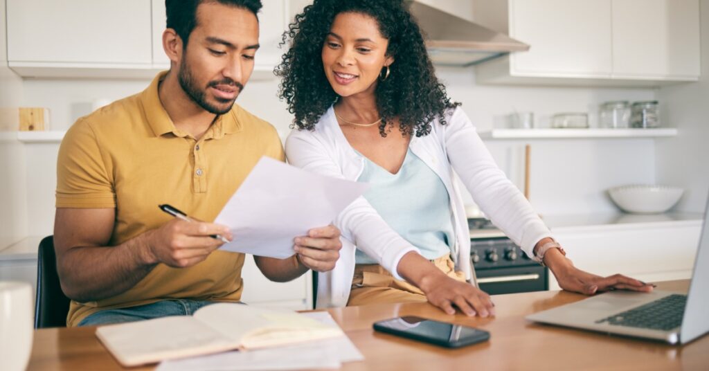 A man and a woman sit together in a bright kitchen and look at a document. Papers and an open laptop are before them.
