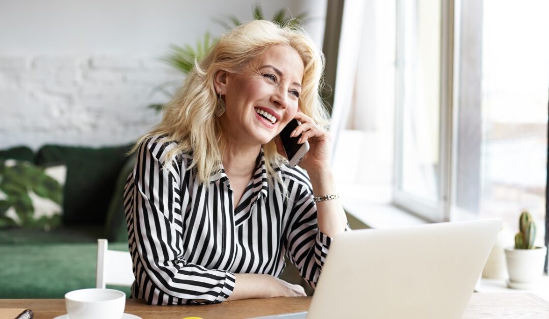 A smiling woman at a table talks on a smartphone. An open laptop, a coffee cup, and a notebook are in front of her.