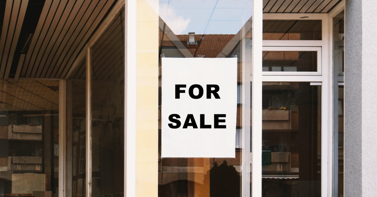 A vacant storefront has large glass windows and a glass entry door. A white sign reading “For Sale” is on a window.