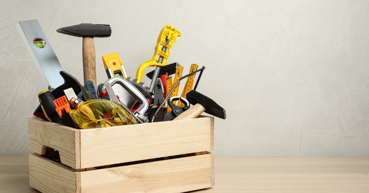 A wooden crate sits on a table. It is full of hammers, levels, rulers, screwdrivers, and other tools and supplies.