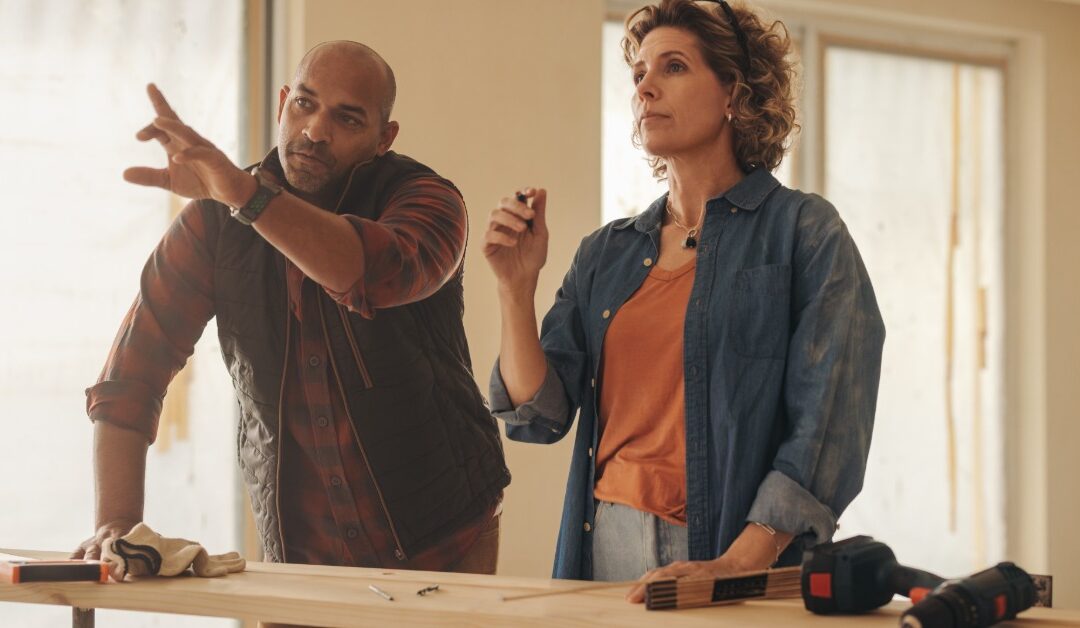 A man and woman stand behind a wooden board inside a partially renovated room. The man points across the space.