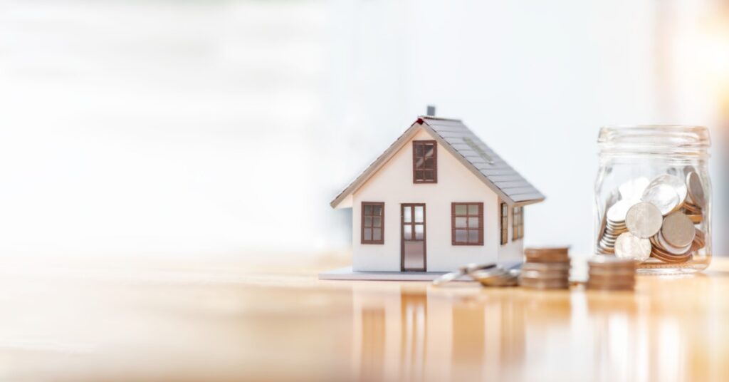 A small model house sits on a reflective surface near stacks of coins. A glass jar filled with coins is to the right.