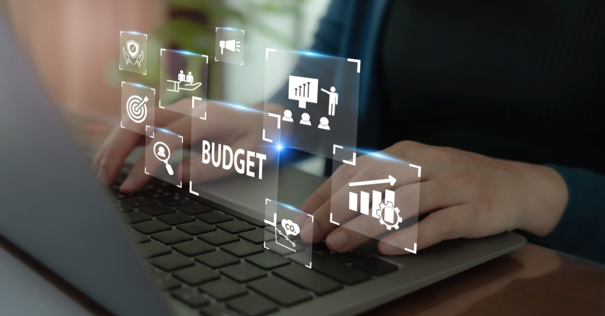A person types on a laptop. Transparent digital icons float above the keyboard, including the word "BUDGET."