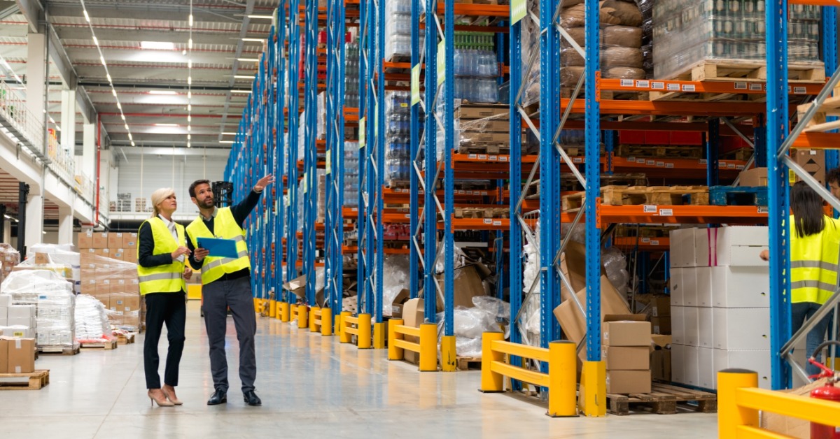 Two people in yellow safety vests stand in a large warehouse aisle beside tall storage racks. One points at the shelves.
