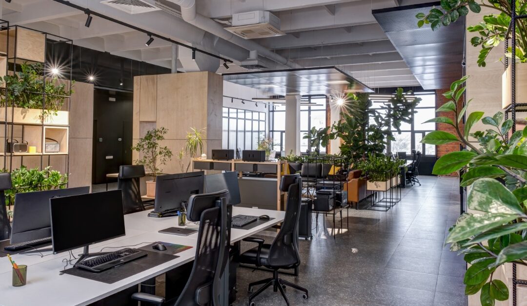 An open-plan office features shared workstations, large plants, wood partitions, and industrial-style ceiling fixtures.