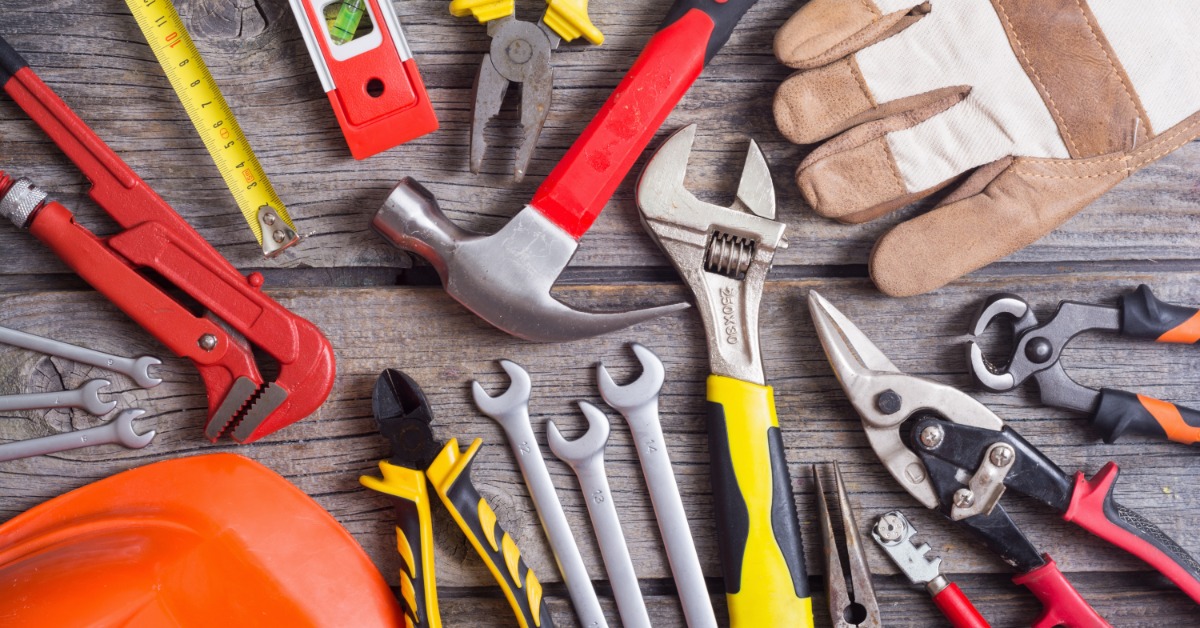 Assorted hand tools are arranged on a wooden surface, including a hammer, wrenches, pliers, a level, and work gloves.