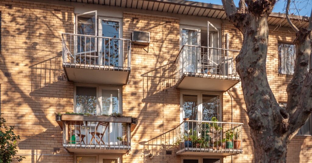 A brick apartment building has several small balconies with metal railings. Two bicycles are by one unit's door.