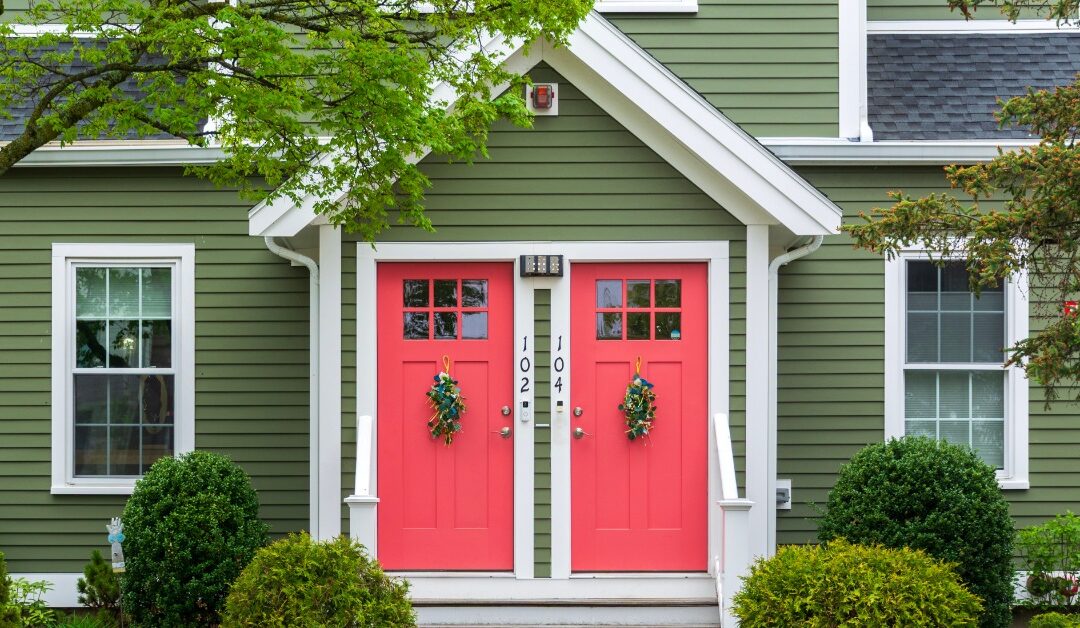 Two coral red front doors sit side by side on a green residential building with white trim. Bushes line the front steps.