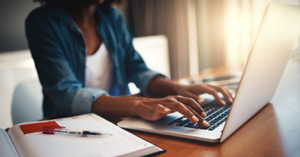 A person types on a laptop as warm sunlight comes through a window. On either side of the laptop are notebooks and pens.