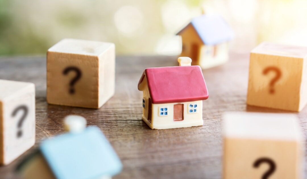Small model houses sit among wooden blocks marked with question marks. A red-roofed house is in focus at the center.