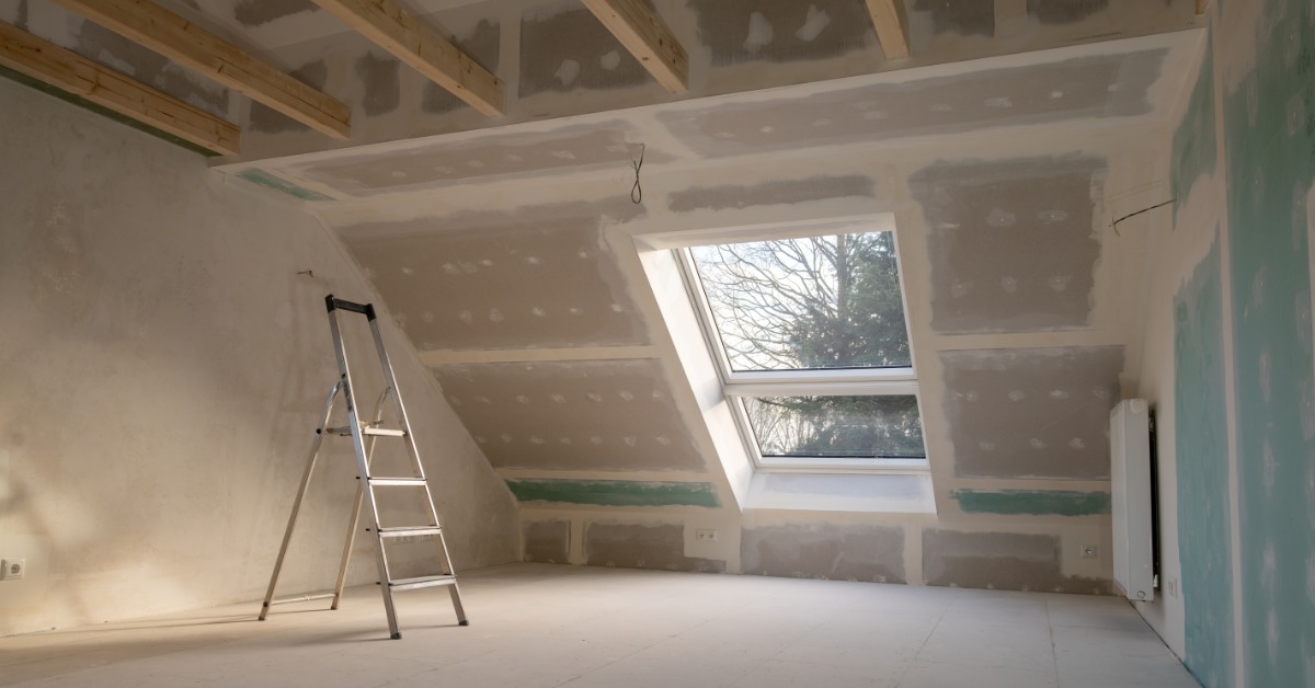An unfinished room has exposed ceiling beams, drywall panels, and a window on a slanted wall. A ladder is near one wall.