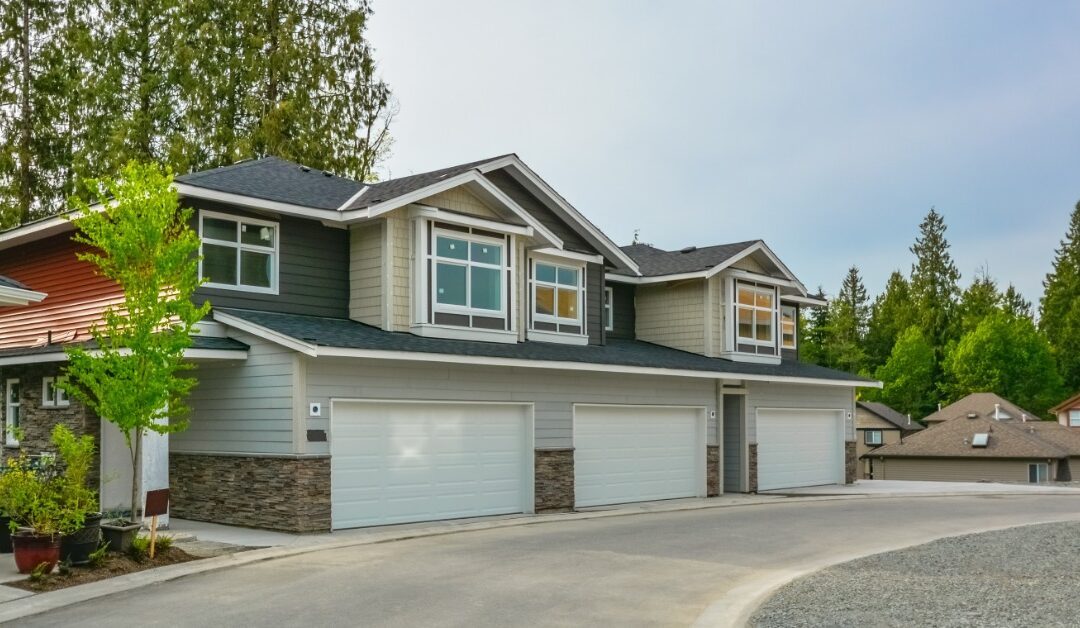 A two-story multifamily building has gray and tan siding, stone accents, and three white double-garage doors.