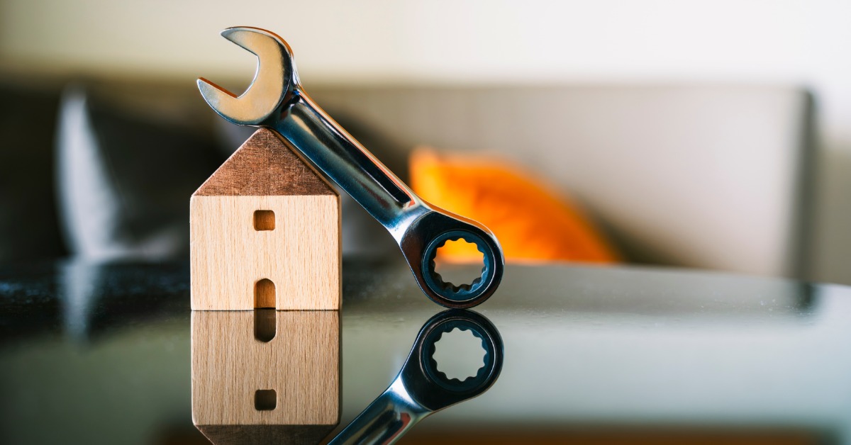 A metal wrench leans on a miniature wooden house on a reflective tabletop. Blurred furnishings are in the background.