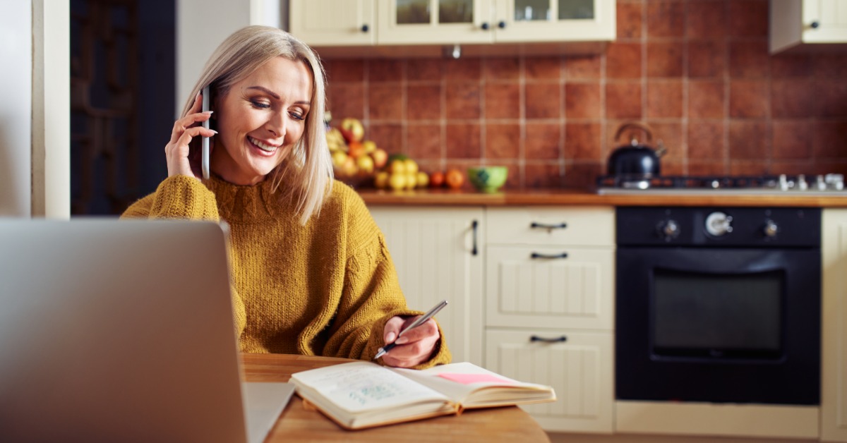 A woman sits at a kitchen table with a laptop and an open notebook. She holds a phone to her ear and writes.