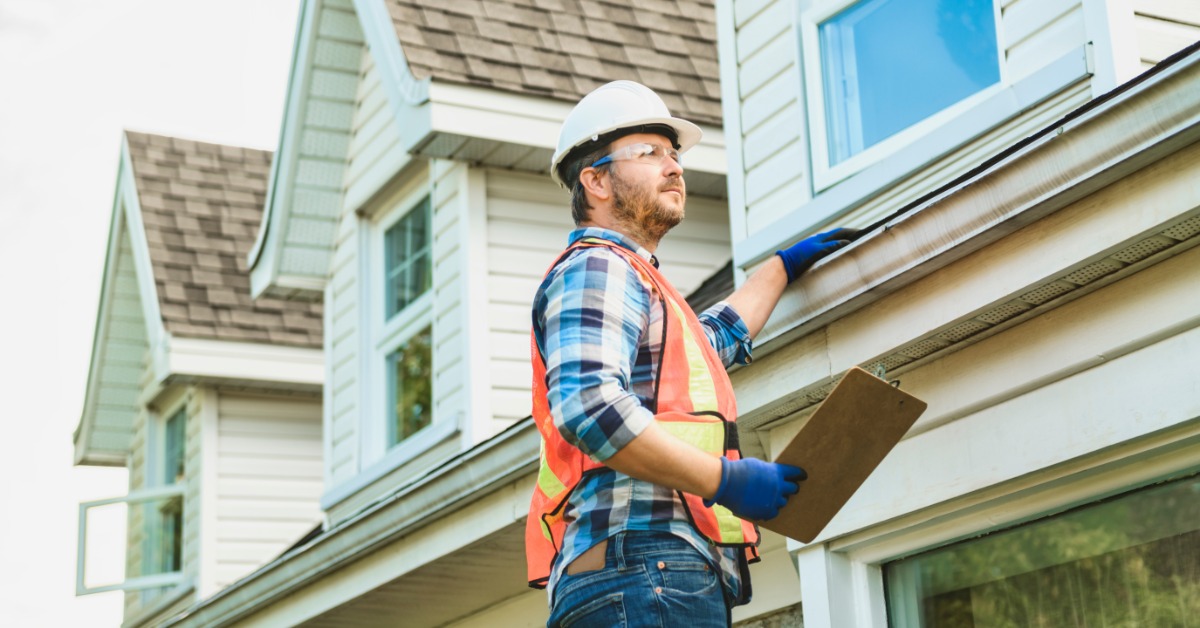 A man in a hard hat and other safety gear touches a roof as he examines it. He holds a clipboard in his other hand.
