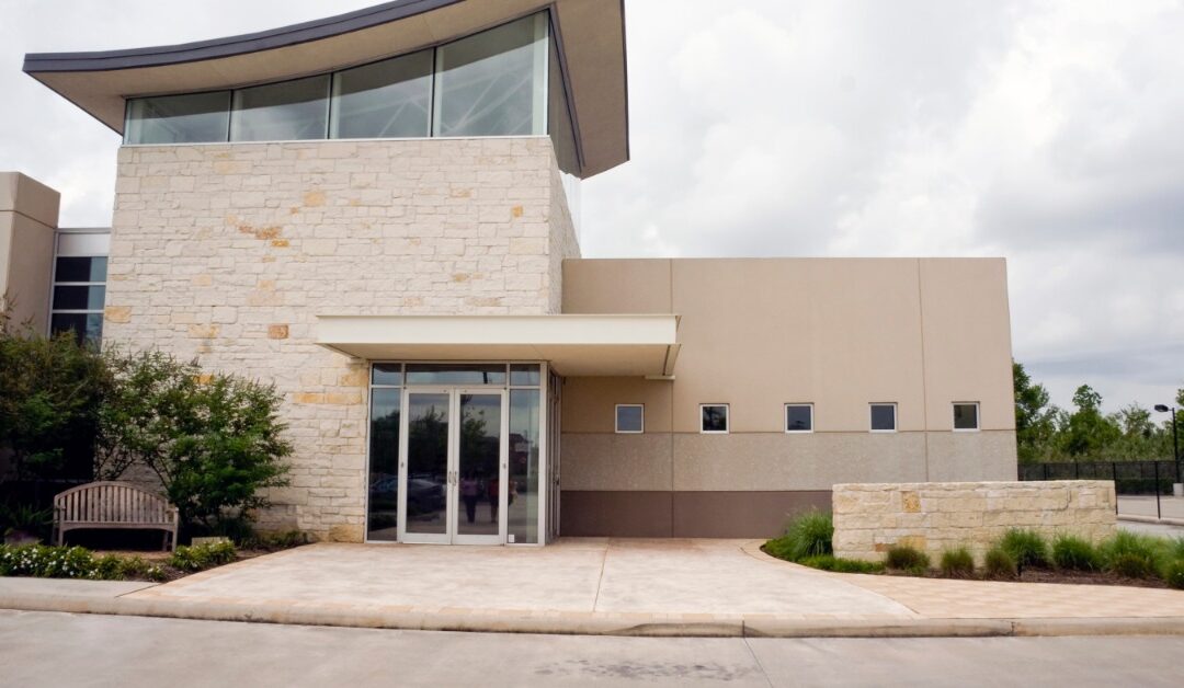 A modern commercial building has a curved roofline, large upper windows, a light stone exterior, and glass double doors.