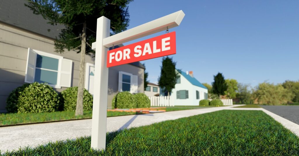 A red "For Sale" sign hangs from a white post in front of a house. A sidewalk, more houses, and a street are nearby.