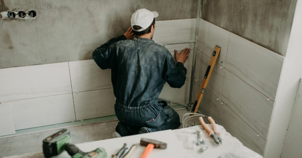 A worker places white, rectangular tiles along an unfinished concrete wall. Hand tools and power tools are nearby.