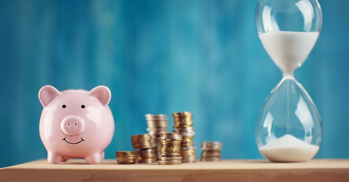 A pink piggy bank, stacks of coins, and a clear glass hourglass filled with white sand are arranged on a table.